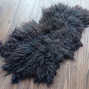 Puede incluir: Una alfombra de piel de oveja gris oscuro con un aspecto lanudo y texturizado. La alfombra tiene forma irregular y est&aacute; sobre un suelo de madera. Las fibras de lana tienen un rizo natural, creando un aspecto suave y acogedor. El color de la alfombra var&iacute;a de gris oscuro a negro.