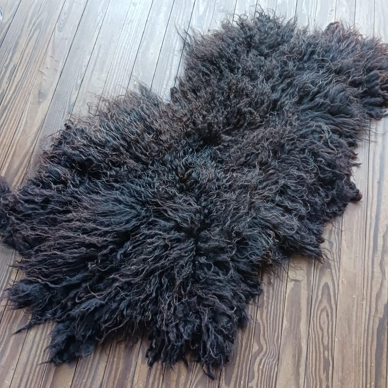 Puede incluir: Una alfombra de piel de oveja gris oscuro con un aspecto lanudo y texturizado. La alfombra tiene forma irregular y est&aacute; sobre un suelo de madera. Las fibras de lana tienen un rizo natural, creando un aspecto suave y acogedor. El color de la alfombra var&iacute;a de gris oscuro a negro.