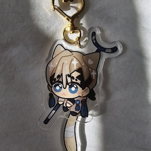 May include: A clear acrylic keychain featuring a cartoon character with cat-like features. The character has light brown hair, blue eyes, and is holding a staff. The keychain has a gold-colored heart-shaped clasp.