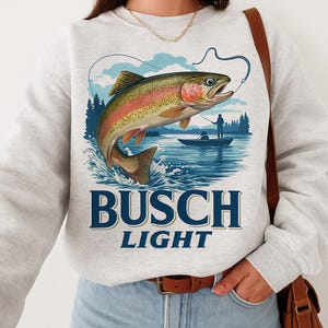 May include: A light gray sweatshirt featuring a colorful illustration of a rainbow trout leaping from water. The design includes a fishing scene with a boat and the words "BUSCH LIGHT" in navy blue.