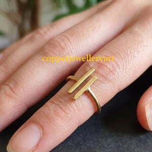 May include: A gold-toned ring featuring two parallel vertical bars on the front. The band is thin and circular, encircling the finger. The text 'copperjewelleryart' is visible in yellow font across the center of the ring design.