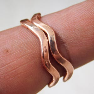 May include: Two copper-colored rings with a wavy design are stacked on a finger. The rings have a smooth, polished finish and a warm, reddish-brown hue. The rings are close together, creating a layered effect.