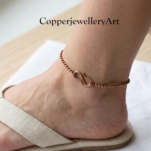 May include: A copper anklet featuring a delicate beaded chain design with a prominent S-shaped hook clasp. The metallic jewelry piece rests around the ankle, showcasing a warm, reddish-brown tone and a simple, functional closure mechanism.