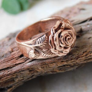 Può includere: Un anello in oro rosa con un design dettagliato di rosa e foglie. Il motivo floreale dell'anello è ben visibile, con dettagli intricati sui petali e sulle foglie. È un gioiello.