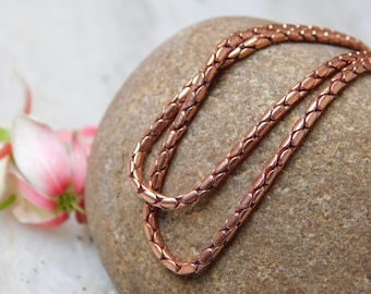 May include: A copper-colored necklace with a textured, linked design rests on a smooth, brown stone. The necklace is curved, showcasing its intricate pattern. A pink and white flower is visible in the background, adding a touch of color.