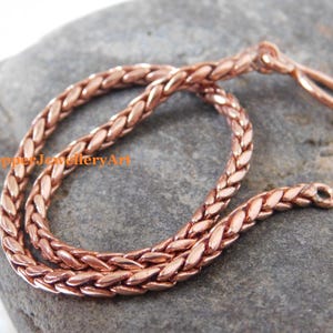 May include: A copper-coloured chain necklace with a woven design. The necklace is made of small, interlocking links, creating a braided appearance. The chain is looped and rests on a grey stone surface. The text "CopperJewelleryArt" is visible.