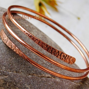 May include: A copper-colored, hammered-texture cuff bracelet. The bracelet wraps around the wrist with a spiral design. The metal has a warm, reddish-brown hue. The bracelet is displayed on a gray stone surface.