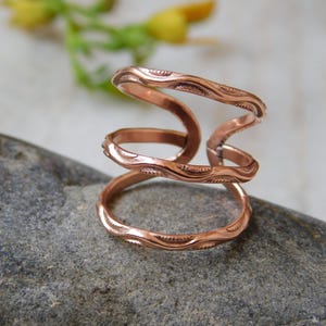 Può includere: Un anello color rame con un design aperto unico. L'anello presenta tre fasce collegate da elementi curvi a forma di onda. Le fasce hanno un motivo strutturato e inciso. L'anello è esposto su una pietra grigia.