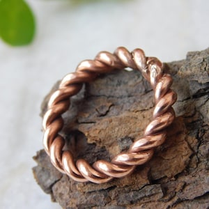 Pode incluir: Um anel cor de cobre com um design de corda torcida. O anel está sobre uma superfície de madeira castanha texturizada. O design do anel apresenta fios entrelaçados, criando uma aparência texturizada. O anel é uma joia.