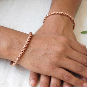 Könnte beinhalten: Zwei Kupferarmbänder mit gedrehtem Seildesign. Jedes Armband ist aus einem einzelnen Strang metallischem Kupferdraht gefertigt, der zu einem durchgehenden Spiralmuster geformt ist und ein strukturiertes, abgerundetes Erscheinungsbild am Handgelenk erzeugt.