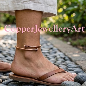 May include: A copper anklet shaped like a bent nail with a metallic finish, featuring a rounded head and a pointed end with small indentations, worn around the ankle above a brown leather flip-flop sandal.