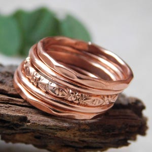 May include: A collection of copper-coloured rings, some with textured and floral patterns. The rings are stacked on a piece of dark brown wood, with a blurred green leaf in the background. The rings have a shiny, metallic finish.