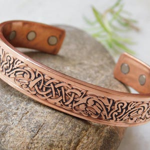 May include: A copper cuff bracelet with an intricate Celtic knot design. The bracelet has a polished finish and is approximately 2.5 inches wide. The design is etched in black, creating a contrast against the copper.