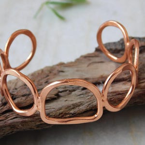 May include: A copper-colored cuff bracelet with an open design. The bracelet features a series of connected, rounded rectangular shapes. The jewelry is displayed on a piece of wood.