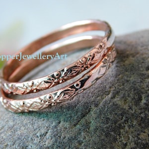 Op de afbeelding: Twee roségouden ringen met een gedetailleerd bloemmotief zijn op elkaar gestapeld. De ringen liggen op een grijze stenen ondergrond. De ringen hebben een glanzende afwerking. De tekst "CopperJewelleryArt" is zichtbaar.