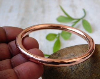 May include: A polished copper bangle bracelet. The round bracelet has a smooth, reflective surface. The copper color is a warm, reddish-brown hue. The bangle is a simple, elegant piece of jewelry.