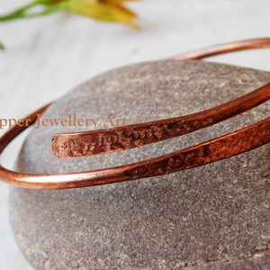 May include: A handcrafted copper bracelet with a hammered texture. The open cuff design wraps around the wrist. The bracelet is displayed on a gray stone surface. The text "Copper Jewellery Art" is visible.