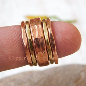 Pode incluir: Um conjunto de anéis de cobre e dourado. Os anéis têm larguras e texturas variadas, com uma faixa de cobre martelado no centro. Os anéis são exibidos em um dedo, destacando o design e o artesanato da joia.