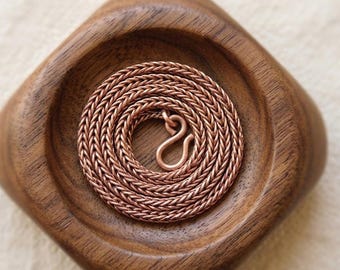 May include: A copper-colored chain necklace with a woven design, coiled inside a wooden bowl. The necklace has a hook clasp. The bowl is made of dark wood and has a square shape with rounded edges.