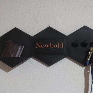 Puede incluir: Un organizador de pared geométrico negro con un estante, ganchos para llaves y el nombre "Newbold" en letras color cobre. Una cartera marrón descansa sobre el estante y las llaves cuelgan de los ganchos.