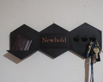 May include: A black, geometric wall-mounted organizer with a shelf, key hooks, and the name "Newbold" in copper-colored lettering. A brown wallet rests on the shelf, and keys hang from the hooks.