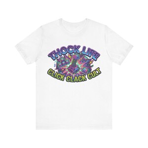 May include: White t-shirt with a vibrant graphic. The design features the text "THOCK LIFE" and "CLICK CLACK CULT", with a central image of a mechanical device. The graphic uses purple, blue, and green colours. A casual, graphic tee.