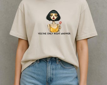 May include: Beige t-shirt featuring a graphic of a woman with dark hair and red lipstick holding a lollipop. The text "YES.THE ONLY RIGHT ANSWER." is printed below the graphic. The shirt is paired with blue denim jeans.