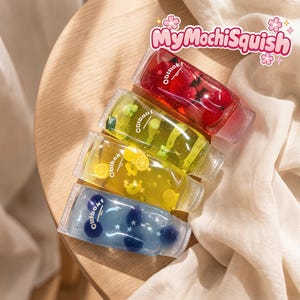 Può includere: Quattro tubetti trasparenti riempiti di gel colorato e piccoli ciondoli a forma di frutta. I tubetti contengono gel rosso, verde lime, giallo e blu con accenti di frutta e stelle. Ogni tubetto riporta la scritta MyMochiSquish in alto.