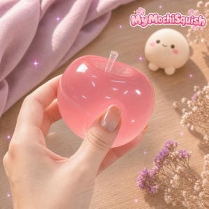 Può includere: Un oggetto traslucido rosa a forma di mela con un gambo trasparente, tenuto in mano. Nelle vicinanze si trova una piccola statuetta rotonda color crema con un viso sorridente e guance rosa, accompagnata dal testo MyMochiSquish nell'angolo in alto a destra.