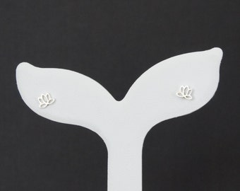 Pode incluir: Um par de brincos de prata com um design de flor de lótus. Os brincos são exibidos em um suporte de brincos branco em forma de cauda de baleia sobre um fundo preto.