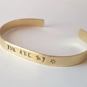 May include: A gold-coloured cuff bracelet with the words "You are my" and a sun symbol stamped on the front. The bracelet has a hammered texture and is open at the ends.