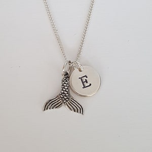 Könnte beinhalten: Silberne Halskette mit einem Meerjungfrauen-Schwanz-Anhänger und einem runden Anhänger mit dem eingravierten Buchstaben E.