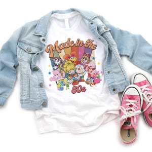 May include: White t-shirt with a colorful graphic featuring cartoon characters and the text "Made in the 80s." A light blue denim jacket and pink sneakers with white laces are also in the image.
