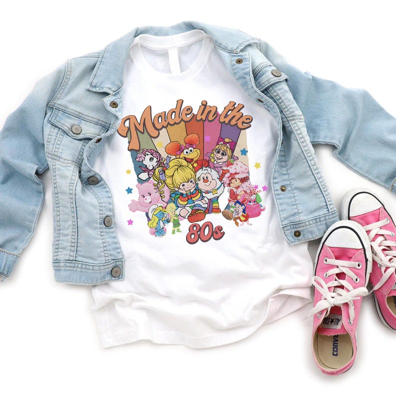 May include: White t-shirt with a colorful graphic featuring cartoon characters and the text "Made in the 80s." A light blue denim jacket and pink sneakers with white laces are also in the image.