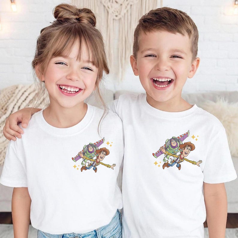 May include: Two children wearing white t-shirts with a colorful graphic of Buzz Lightyear and Woody from Toy Story. The graphic shows the characters flying. The children are smiling.