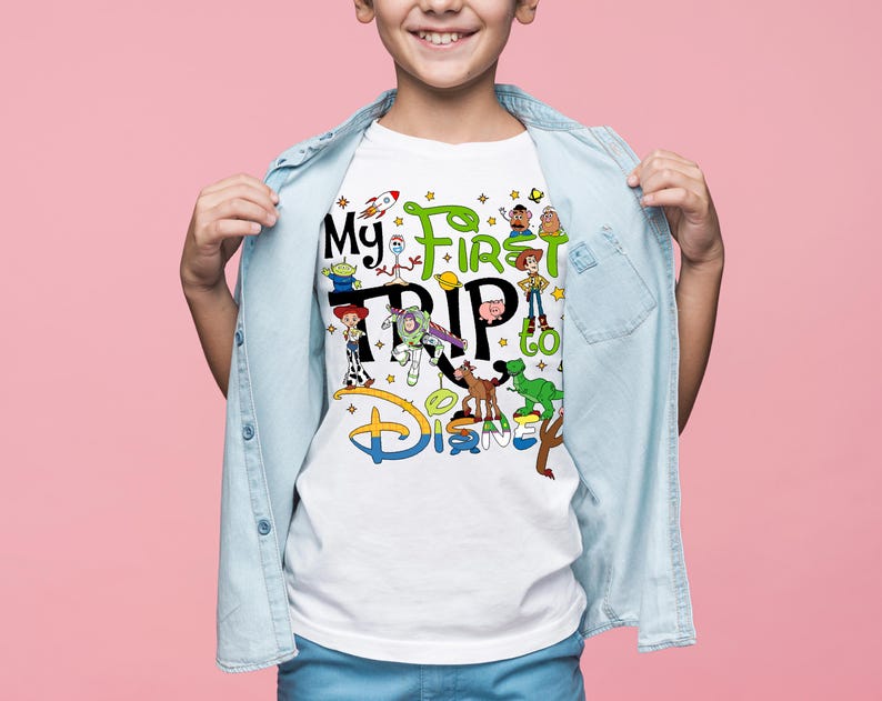 May include: White t-shirt with the text "My First Trip to Disney" and various cartoon characters. The design includes a rocket, stars, and the Disney logo in blue and yellow. The shirt is worn by a child.
