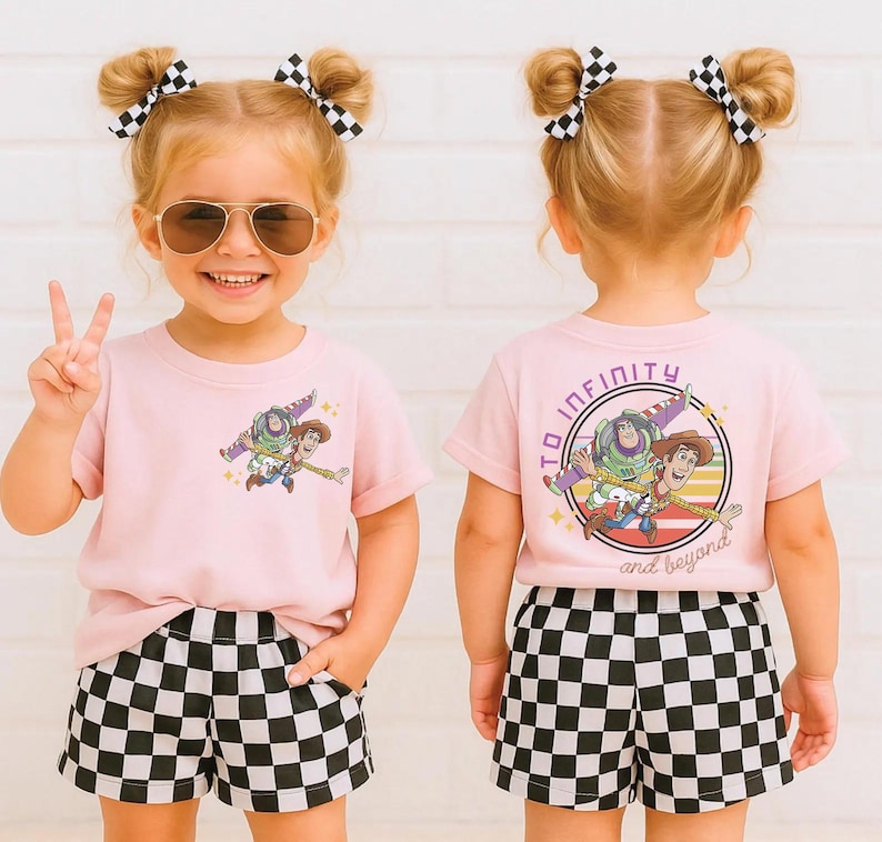 May include: A young child wearing a light pink t-shirt and black and white checkered shorts. The t-shirt features a graphic of Buzz Lightyear and Woody from Toy Story. The text "To Infinity and Beyond" is on the back of the shirt.