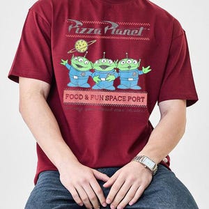 Puede incluir: Camiseta burdeos con el logo de Pizza Planet y tres extraterrestres verdes con trajes espaciales azules. La camiseta tambi&eacute;n tiene el texto "FOOD & FUN SPACE PORT". La camiseta es de manga corta y cuello redondo.
