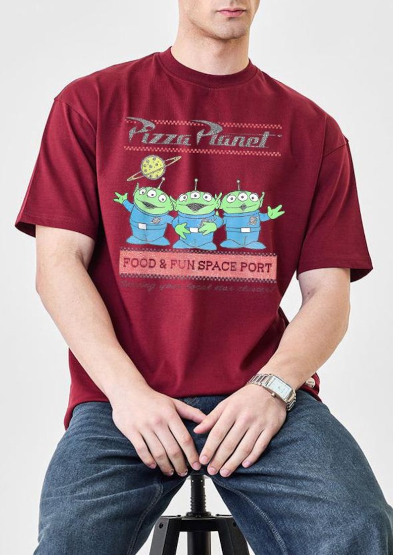Puede incluir: Camiseta burdeos con el logo de Pizza Planet y tres extraterrestres verdes con trajes espaciales azules. La camiseta tambi&eacute;n tiene el texto "FOOD & FUN SPACE PORT". La camiseta es de manga corta y cuello redondo.