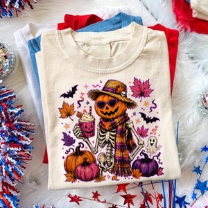 Peut inclure: Un t-shirt couleur crème avec un motif représentant un squelette portant un chapeau de paille, des lunettes de soleil en forme de cœur et une écharpe à carreaux. Il tient une tasse citrouille entouré de citrouilles, chauves-souris et feuilles.