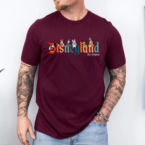 Pu&ograve; includere: T-shirt bordeaux con la scritta "Disneyland" in lettere colorate e personaggi dei cartoni animati. Sotto, il testo "The Original". La maglietta &egrave; a girocollo e maniche corte.