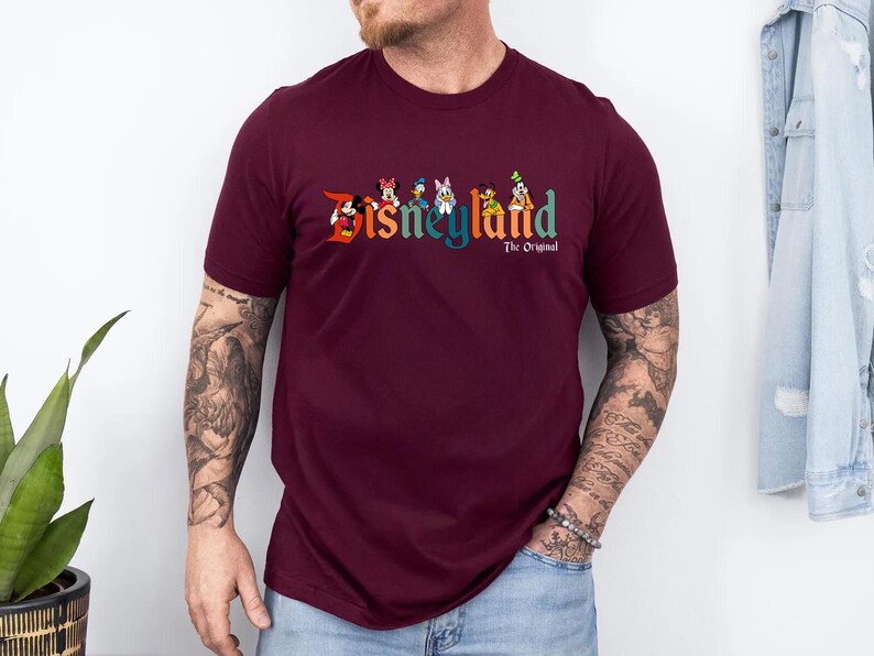 Pu&ograve; includere: T-shirt bordeaux con la scritta "Disneyland" in lettere colorate e personaggi dei cartoni animati. Sotto, il testo "The Original". La maglietta &egrave; a girocollo e maniche corte.