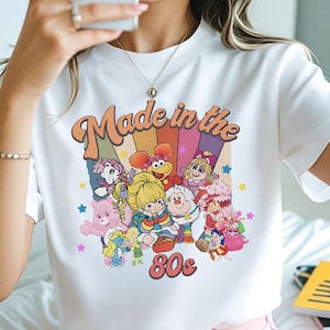 May include: White t-shirt with a colorful graphic featuring cartoon characters and the text "Made in the 80s." The design includes a rainbow background and the number "80s" in a retro font. The shirt has short sleeves.