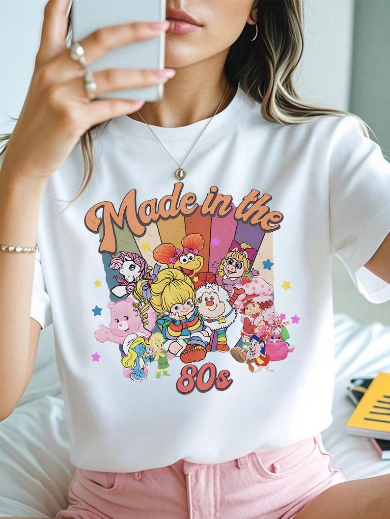 May include: White t-shirt with a colorful graphic featuring cartoon characters and the text "Made in the 80s." The design includes a rainbow background and the number "80s" in a retro font. The shirt has short sleeves.