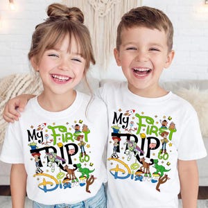 May include: Two children wearing white t-shirts with a colorful graphic. The shirts say "My First Trip to Disney" with cartoon characters. The design includes a rocket, stars, and the Disney logo.