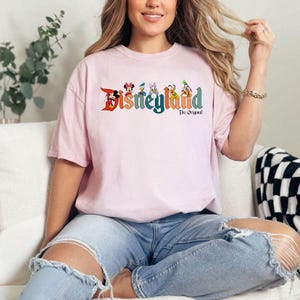 Pu&ograve; includere: Una maglietta rosa chiaro con la scritta "Disneyland" in lettere colorate e personaggi dei cartoni animati. Il testo "The Original" &egrave; sotto la parola "Disneyland". La maglietta &egrave; indossata da una persona che indossa jeans blu.