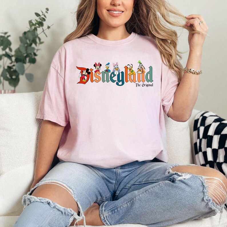 Pu&ograve; includere: Una maglietta rosa chiaro con la scritta "Disneyland" in lettere colorate e personaggi dei cartoni animati. Il testo "The Original" &egrave; sotto la parola "Disneyland". La maglietta &egrave; indossata da una persona che indossa jeans blu.