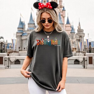 Pu&ograve; includere: T-shirt grigio scuro con il logo Disneyland in testo colorato e personaggi dei cartoni animati. La persona indossa un cerchietto con orecchie di Minnie con fiocco a pois rossi e occhiali da sole. Sullo sfondo c'&egrave; un castello.