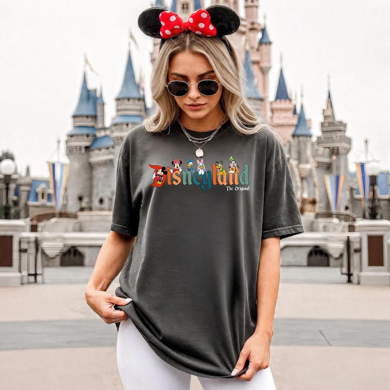 Pu&ograve; includere: T-shirt grigio scuro con il logo Disneyland in testo colorato e personaggi dei cartoni animati. La persona indossa un cerchietto con orecchie di Minnie con fiocco a pois rossi e occhiali da sole. Sullo sfondo c'&egrave; un castello.