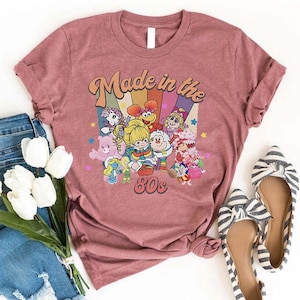 May include: A dusty rose t-shirt featuring a colorful graphic of cartoon characters with the text "Made in the 80s". The design includes a rainbow background. A pair of striped flats and a bouquet of white flowers are also in the image.